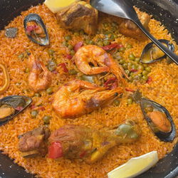 Ein Bild, dass Paella, ein traditionelles Gericht aus Reis, Muscheln, Garnelen, Gemüse und Hühnerfleisch in einer Pfanne zubereitet zeigt